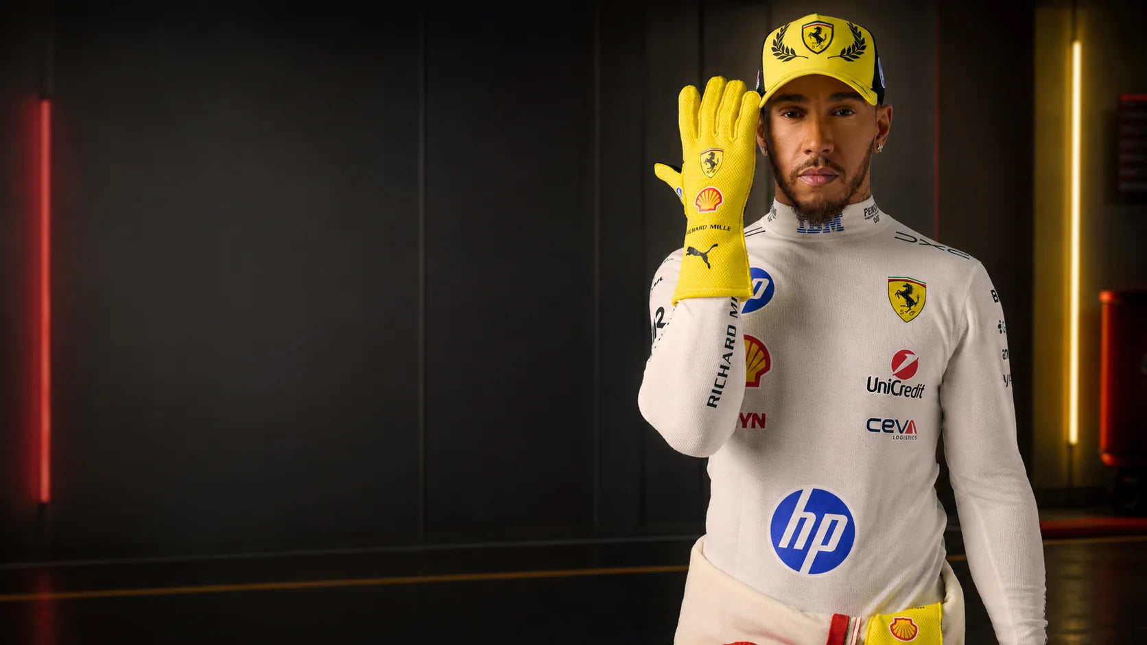 Formula racing driver in a white suit raising a yellow-gloved hand in a dark motorsport garage